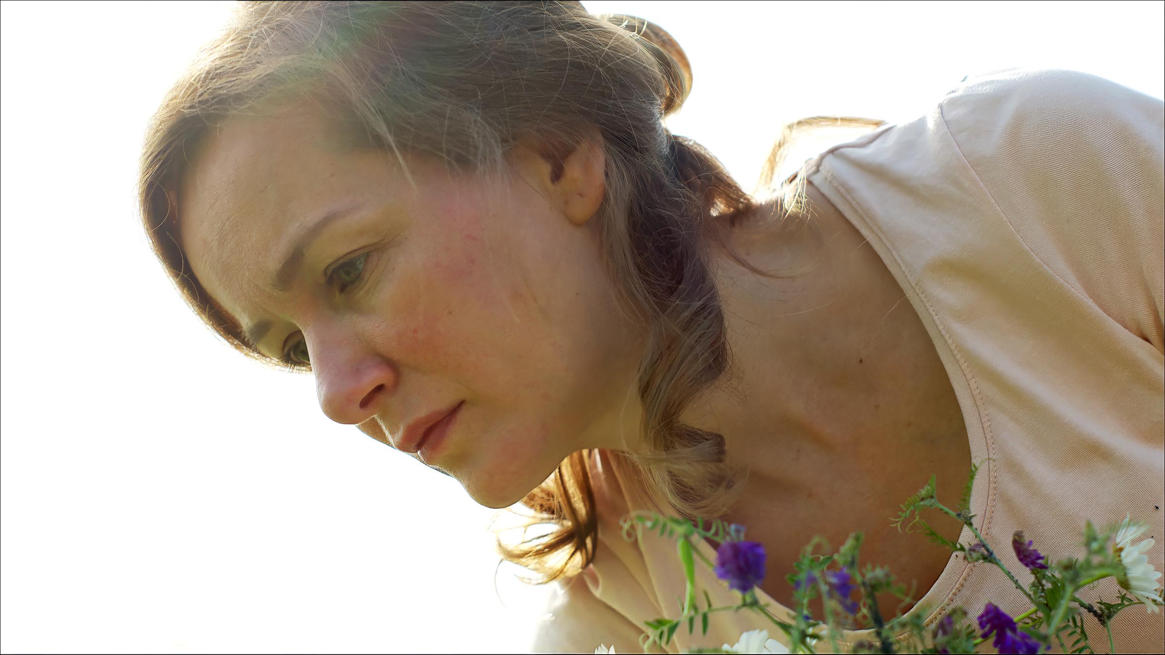 Zana (Adriana Matoshi), eine junge Frau mit langem lockigem Haar, schaut traurig zu Boden. Im Vordergrund sieht man farbige Sommerblüten.