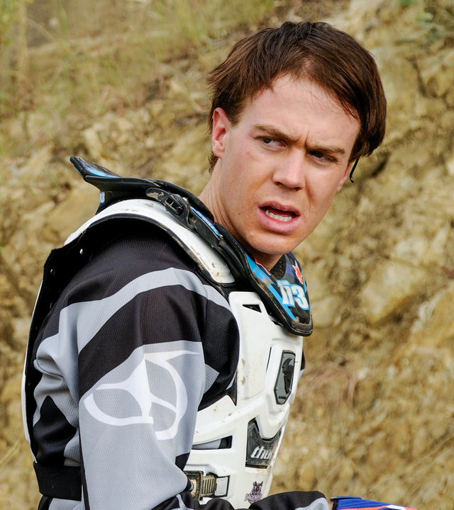 Zane und Nate tragen ihre Motocross-Outfits und stehen an der Startlinie für das Rennen. Zane schaut Nate mit einem ernsten Blick an, dieser lächelt etwas.