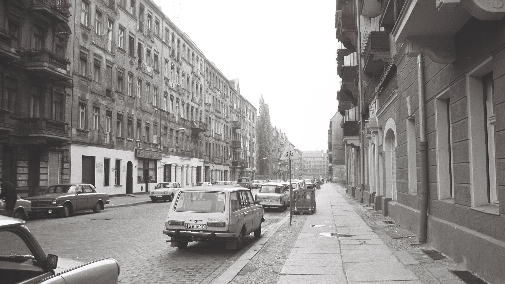 "Mein Land will nicht verschwinden": Schwarzweißfotografie einer städtischen Straßenszene in Ost-Berlin: Ende der 1980er Jahre. Links und rechts der Straße stehen mehrstöckige Altbauten, deren Fassaden teilweise verwittert wirken, mit bröckelndem Putz und Spuren von Abnutzung versehen sind. Die Straße ist mit Kopfsteinpflaster belegt, am rechten Rand verläuft ein breiter Gehweg mit großen Betonplatten auf denen sich Pfützen gebildet haben. Mehrere Automobile der Fabrikanten Trabant und Wartburg parken beidseitig entlang der Straße.