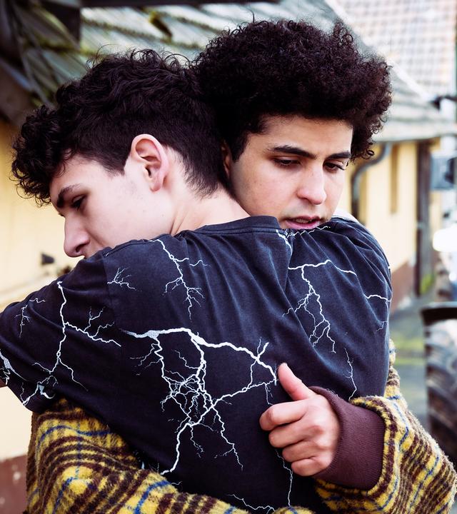 „Von uns wird es keiner sein“: Vor einem Gebäude: Erik (Giuseppe Bonvissuto) umarmt Waldi (Lukas von Horbatschewsky). Im Hintergrund steht ein Traktor.