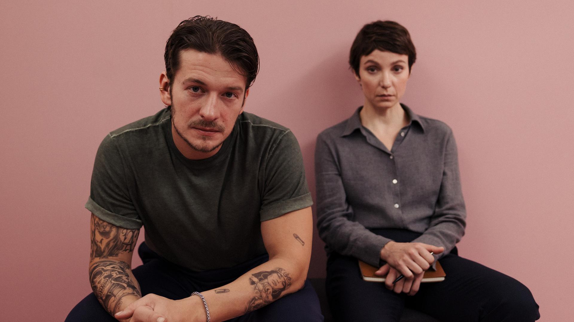 "Im Schatten der Angst - Der Skorpion": Anton Lisky (Stefan Gorski) und Karla Eckhardt (Julia Koschitz) sitzen nebeneinander auf einer schlichten Bank im mobiliarlosen "Pink Room", einem ganz in Rosa gehaltenen Raum. Lisky hat sich vorgebeugt, die Hände ineinandergelegt. Seine Arme sind mit Tätowierungen geschmückt.