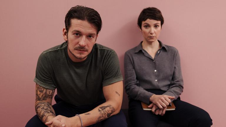 "Im Schatten der Angst - Der Skorpion": Anton Lisky (Stefan Gorski) und Karla Eckhardt (Julia Koschitz) sitzen nebeneinander auf einer schlichten Bank im mobiliarlosen "Pink Room", einem ganz in Rosa gehaltenen Raum. Lisky hat sich vorgebeugt, die Hände ineinandergelegt. Seine Arme sind mit Tätowierungen geschmückt.