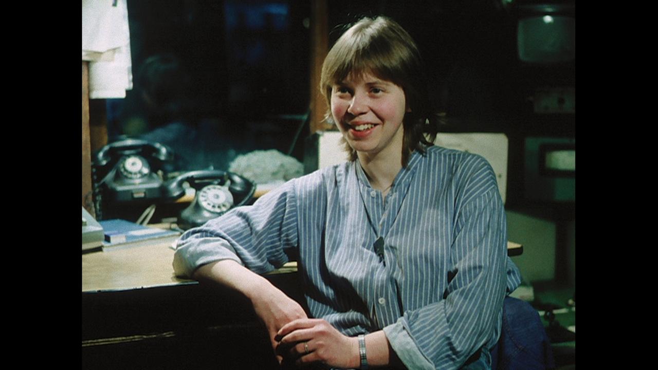 "Die Unbeugsamen 2 – Guten Morgen, ihr Schönen": Ein Bild aus einem Film aus den 1980er-Jahren. Eine junge Frau mit kurzen Haaren sitzt in Arbeitskleidung an einem Tisch. Im Hintergrund sind Telefone und Arbeitsgeräte zu sehen.