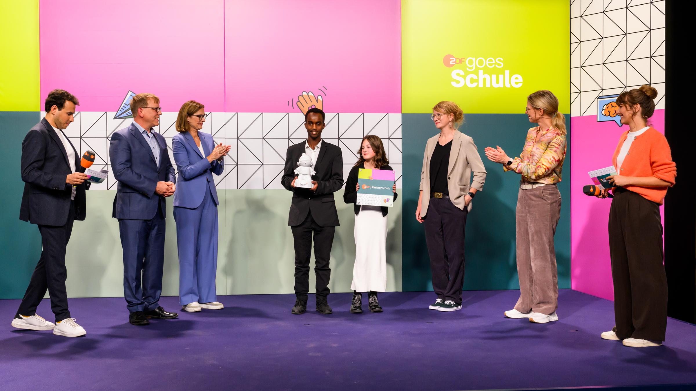 Ein Schüler und eine Schülerin der IGS Wilhelmshaven mit einer Austzeichung von "ZDF goes Schule" vor dem Kampagnenlogo mit ZDF-Moderatorinnen und -Moderatoren und den applaudierenden ZDF-Chefredakteurin Bettina Schausten und Programmdirektorin Nadine Bilke