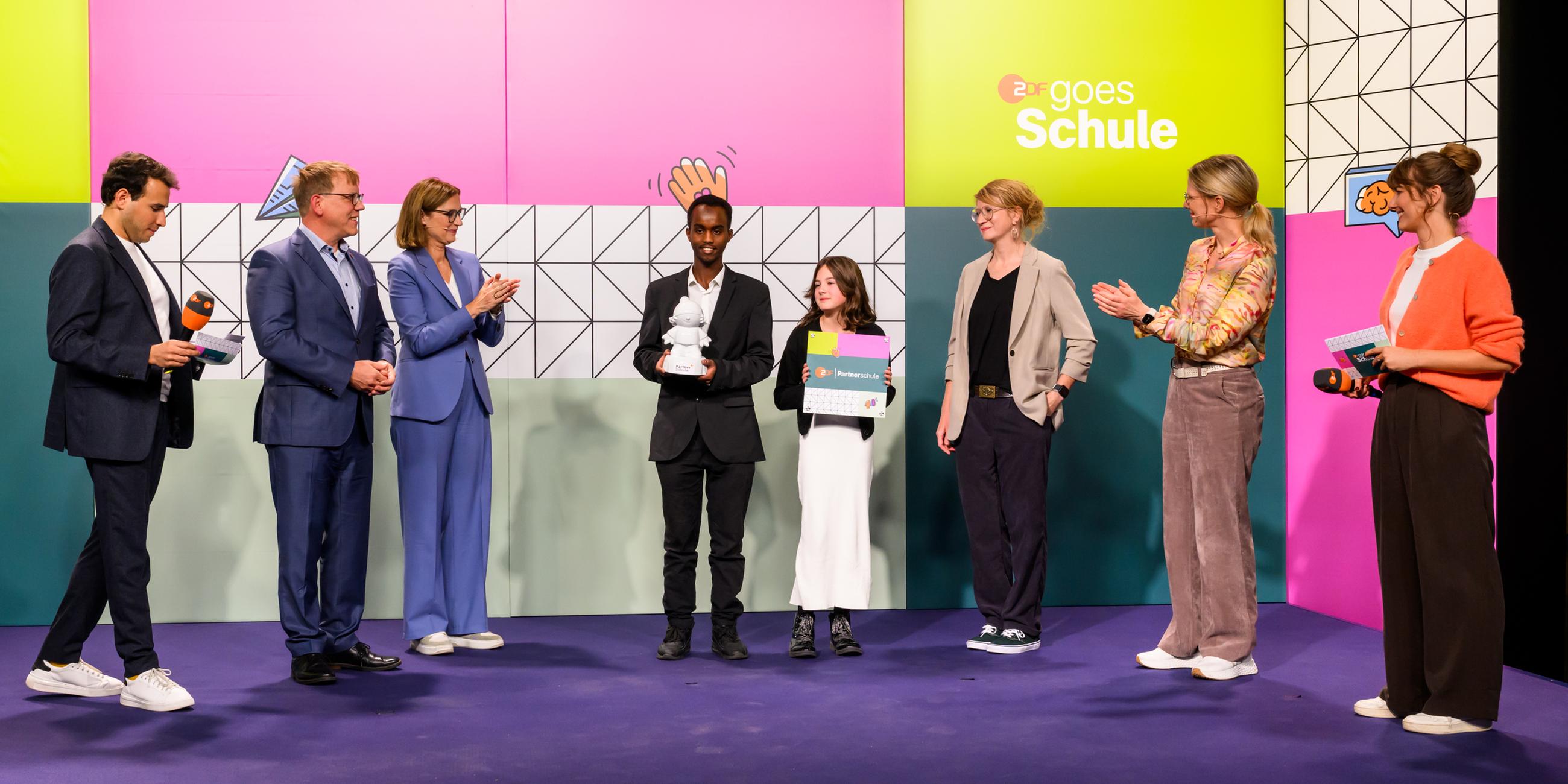 Ein Schüler und eine Schülerin der IGS Wilhelmshaven mit einer Austzeichung von "ZDF goes Schule" vor dem Kampagnenlogo mit ZDF-Moderatorinnen und -Moderatoren und den applaudierenden ZDF-Chefredakteurin Bettina Schausten und Programmdirektorin Nadine Bilke
