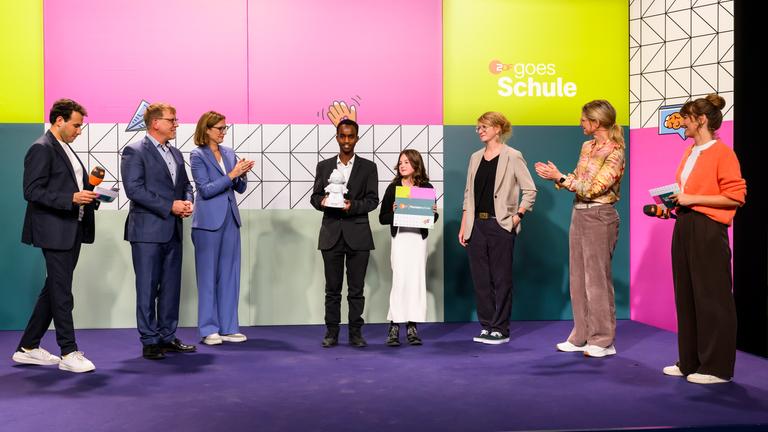 Ein Schüler und eine Schülerin der IGS Wilhelmshaven mit einer Austzeichung von "ZDF goes Schule" vor dem Kampagnenlogo mit ZDF-Moderatorinnen und -Moderatoren und den applaudierenden ZDF-Chefredakteurin Bettina Schausten und Programmdirektorin Nadine Bilke
