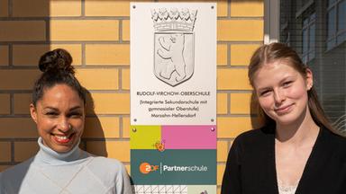 ZDF-Moderatorin und „ZDF goes Schule“-Botschafterin Jana Pareigis steht neben Schülersprecherin Hannah Stephan vor einer Backsteinwand. Zwischen ihnen ist ein Schild der Rudolf-Virchow-Oberschule in Berlin angebracht, beide blicken lächelnd in die Kamera