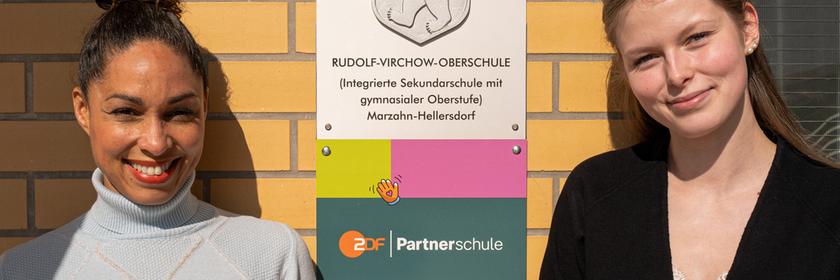 ZDF-Moderatorin und „ZDF goes Schule“-Botschafterin Jana Pareigis steht neben Schülersprecherin Hannah Stephan vor einer Backsteinwand. Zwischen ihnen ist ein Schild der Rudolf-Virchow-Oberschule in Berlin angebracht, beide blicken lächelnd in die Kamera