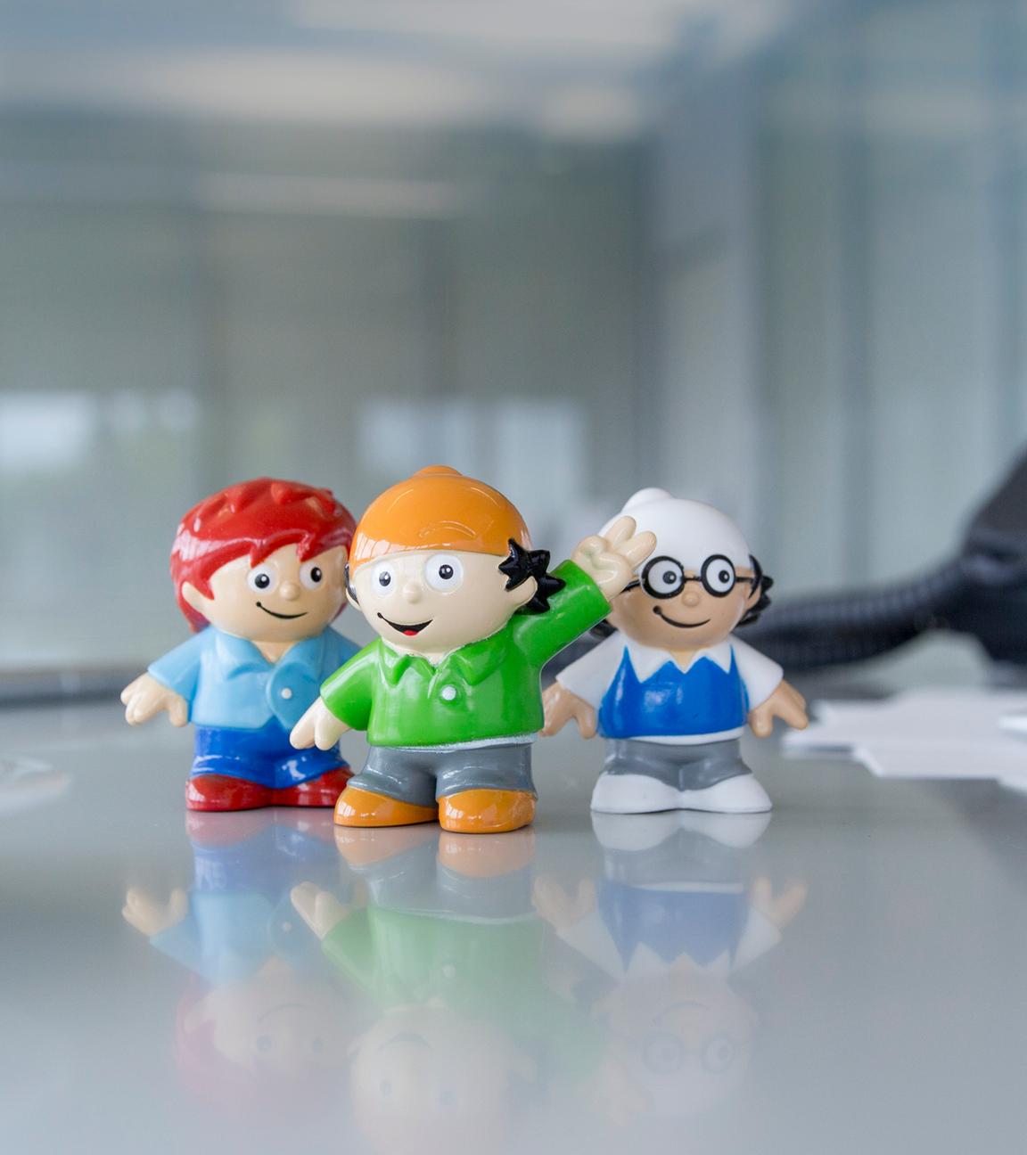 drei Mainzelmännchen-Figuren auf einem Schreibtisch. Links eine Kafeetasse, rechts ein Tischtelefon