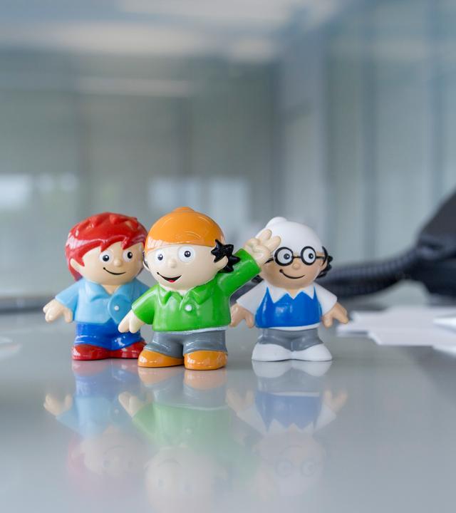 drei Mainzelmännchen-Figuren auf einem Schreibtisch. Links eine Kafeetasse, rechts ein Tischtelefon