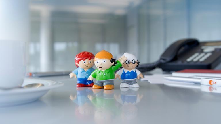 drei Mainzelmännchen-Figuren auf einem Schreibtisch. Links eine Kafeetasse, rechts ein Tischtelefon