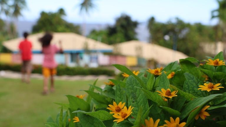 Kinderhilfe Philippinen: Im Vordergrunf gelbe Blumen; im unscharfen Hintergrund laufen zwei Kinder auf ein Haus zu