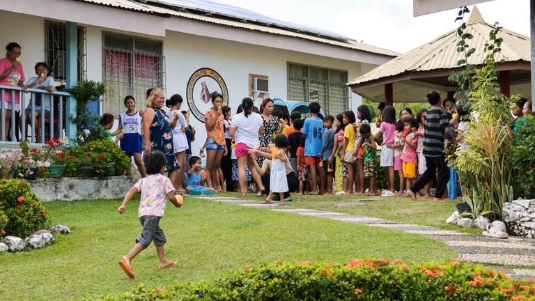 Kinderhilfe Philippinen: Vor einem Haus eine Schlange von Kindern, die auf ein Eis warten, dass eine Frau austeilt. Im Vordergrund läuft ein kleiner Junge auf die Schlange zu