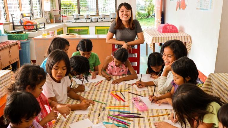 Kinderhilfe Philippinen: Zwölf Kinder sitzen an einem Tisch und basteln Postkarten. Dahinter stützt sich eine junge Frau mit en Armen auf eine Stuhlehen und lächelt in die Kamera