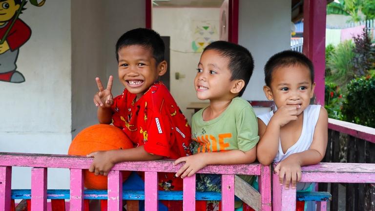 Kinderhilfe Philippinen: Drei kleine Jungen blicken fröhlich über den Zaun einer Veranda