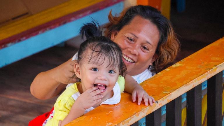 Kinderhilfe Philippinen: Ein kleines Mädchen stützt sich auf das Geländer einer Terasse und wird dabei von der Hausmutter gehalten