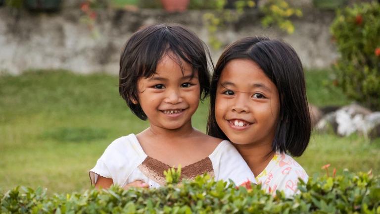 Kinderhilfe Philippinen: Zwei Mädchen blicken lächelnd über eine Hecke