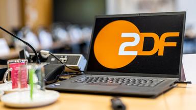 Ein Laptop mit groß dargestelltem ZDF-Logo steht auf einem Konferenztisch während einer Sitzung des ZDF-Verwaltungsrats. Im Vordergrund sind Mikrofon, Getränke und Unterlagen zu sehen, im Hintergrund weitere Sitzplätze unscharf erkennbar.