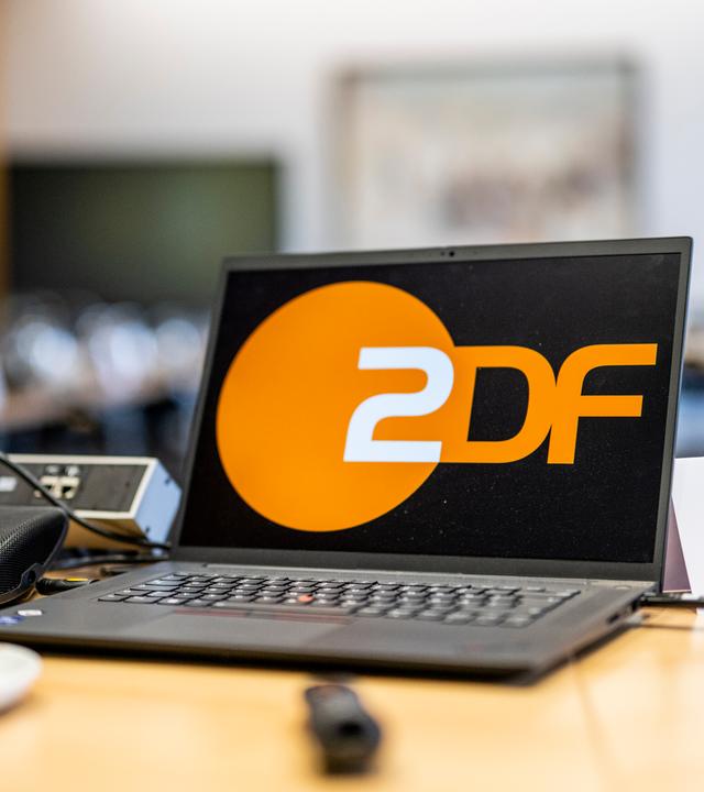 Ein Laptop mit groß dargestelltem ZDF-Logo steht auf einem Konferenztisch während einer Sitzung des ZDF-Verwaltungsrats. Im Vordergrund sind Mikrofon, Getränke und Unterlagen zu sehen, im Hintergrund weitere Sitzplätze unscharf erkennbar.