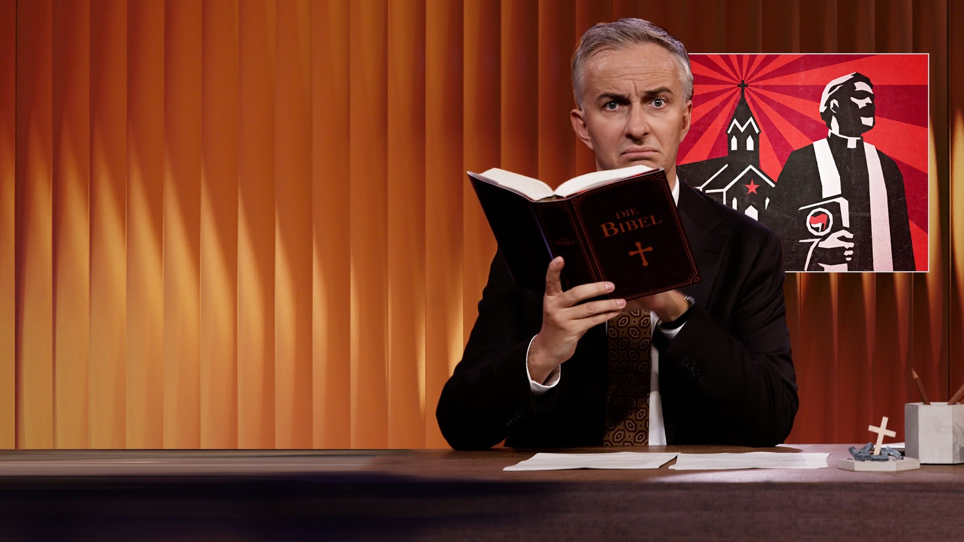 Jan Böhmermann sitzt am Schreibtisch. Er hält eine Bibel. Rechts von ihm ist die Illustration eines Priesters vor einer Kirche zu sehen.