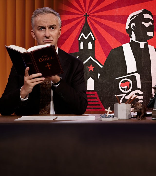 Jan Böhmermann sitzt am Schreibtisch. Er hält eine Bibel. Rechts von ihm ist die Illustration eines Priesters vor einer Kirche zu sehen.