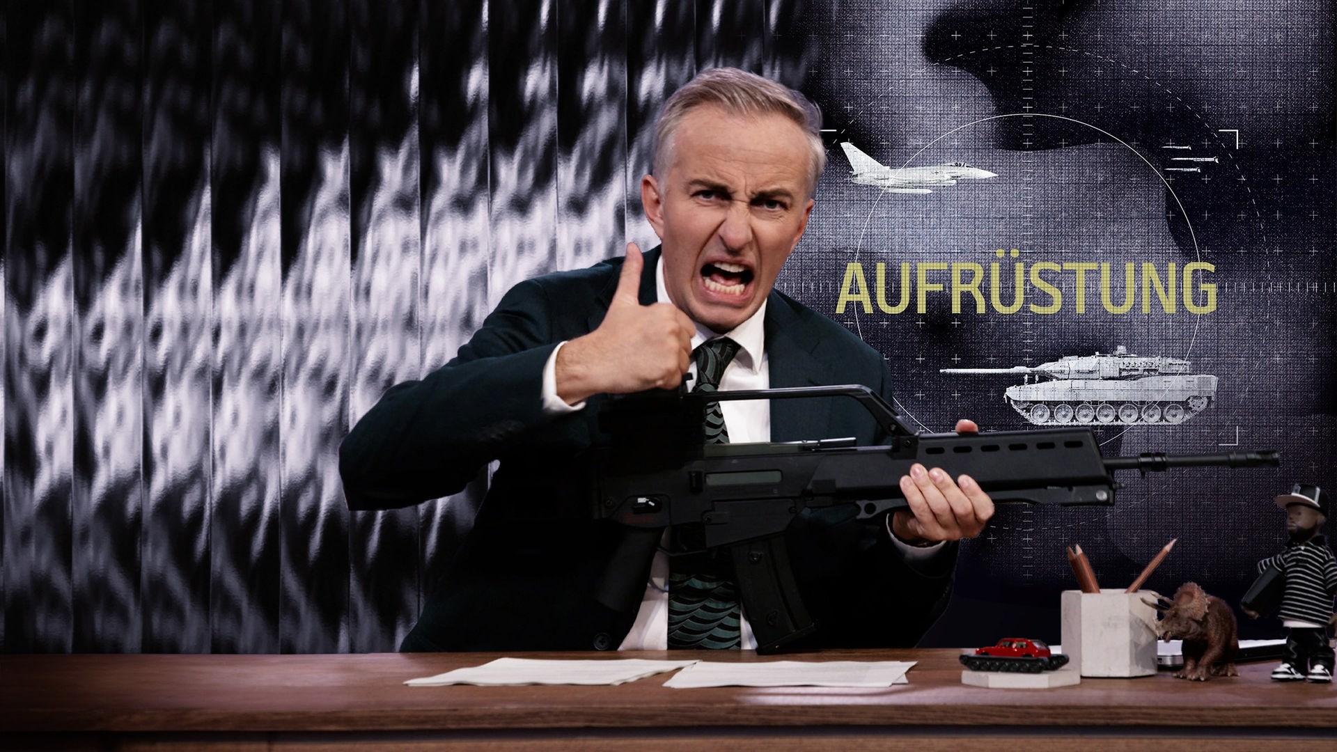 Jan Böhmermann sitzt am Schreibtisch. Er hält eine schwarze Waffe. Rechts im Bild steht das Wort "Aufrüstung". Darüber ist ein Kampfflugzeug und darunter ein Panzer abgebildet.