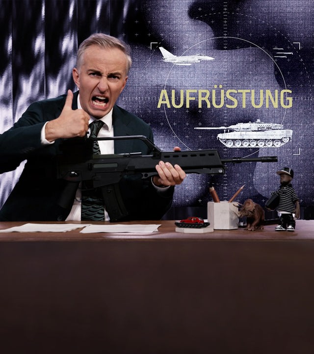 Jan Böhmermann sitzt am Schreibtisch. Er hält eine schwarze Waffe. Rechts im Bild steht das Wort "Aufrüstung". Darüber ist ein Kampfflugzeug und darunter ein Panzer abgebildet.