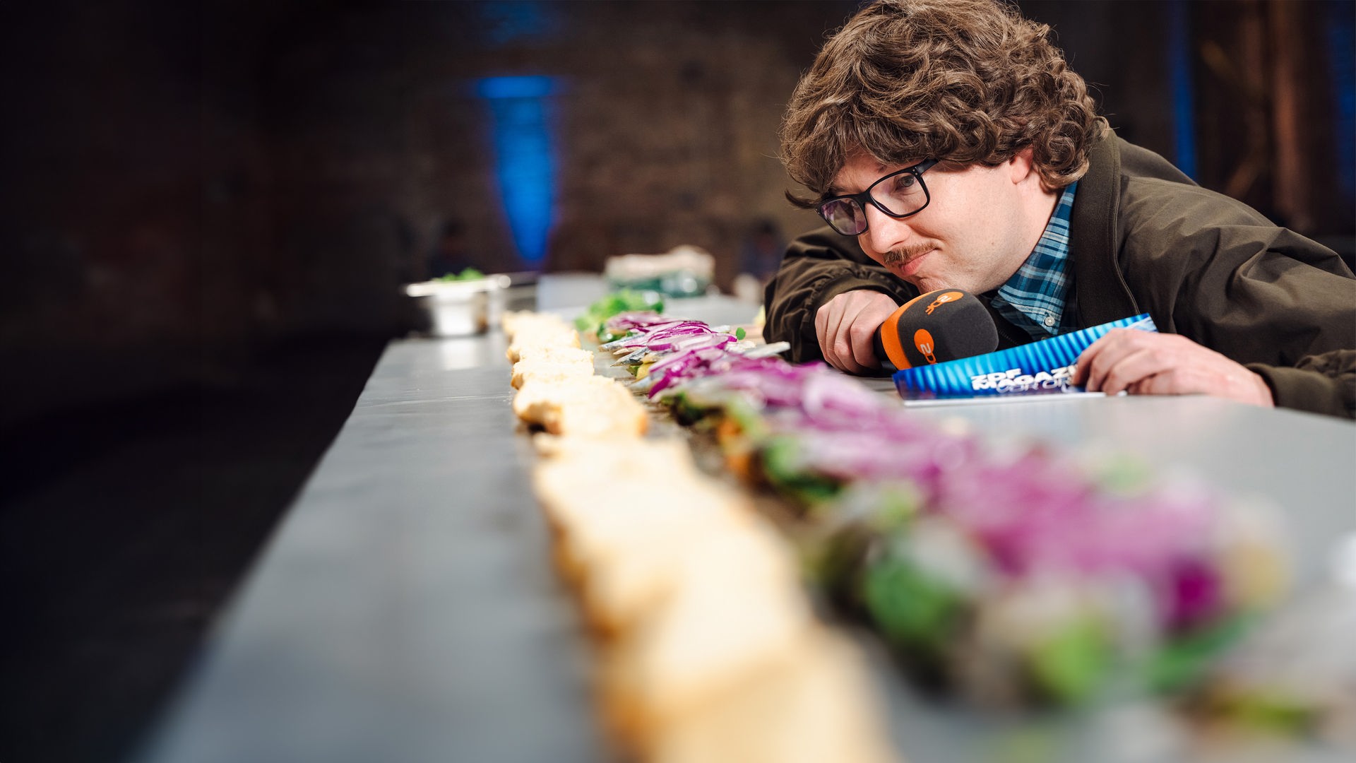 "ZDF Magazin vor Ort": Peter Stefan Thomas betrachtet das längste Matjesbrötchen der Welt. In der Hand hält er ein Mikrofon und Moderationskarten.