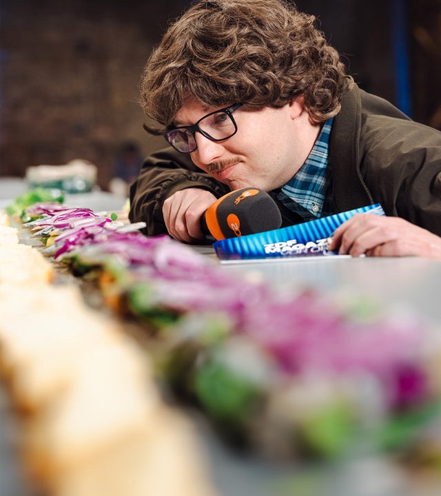 "ZDF Magazin vor Ort": Peter Stefan Thomas betrachtet das längste Matjesbrötchen der Welt. In der Hand hält er ein Mikrofon und Moderationskarten.