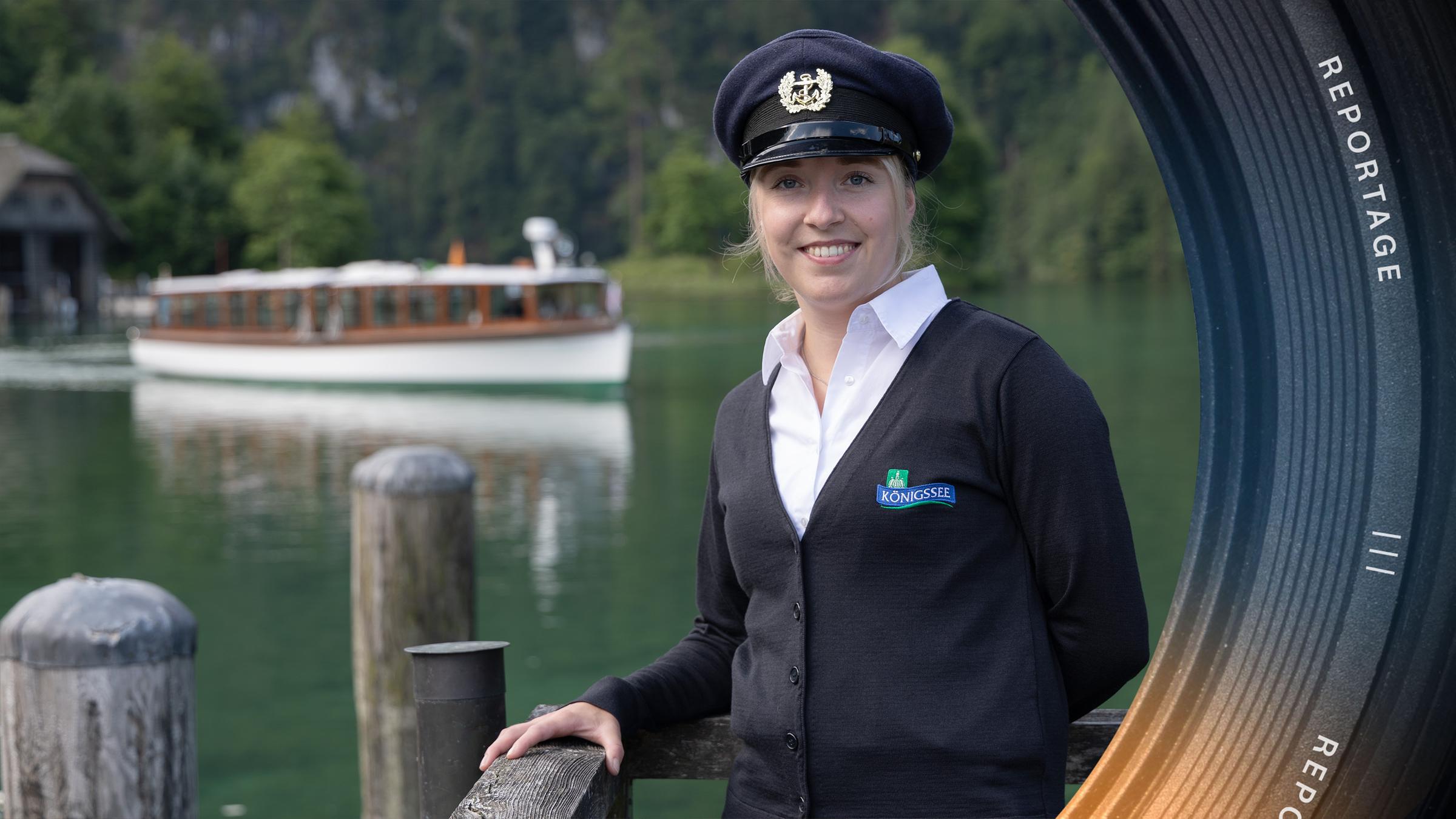 Kapitänin Lena trägt eine Kapitänsmütze, steht am Ufer an einer Holzbefestigung und schaut lächelnd in die Kamera. Im Hintergrund fährt ein Schiff über den Königssee.