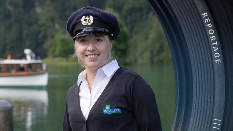 Kapitänin Lena trägt eine Kapitänsmütze, steht am Ufer an einer Holzbefestigung und schaut lächelnd in die Kamera. Im Hintergrund fährt ein Schiff über den Königssee.