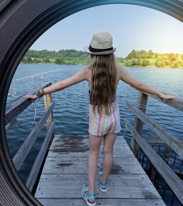 Ein junges Mädchen steht mit dem Rücken zur Kamera auf einem Holzsteg am Gemündener Maar in der Eifel.