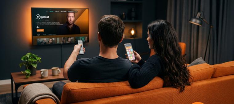 KI-generiertes Bild zu ZDF-Inhalten bei joyn: Ein Pärchen mit Fernbedienungen auf einer orangenen Couch. An der Wand hängt ein Fernseher mit Sven Voss und dem Logo von Aktenzeichen XY
