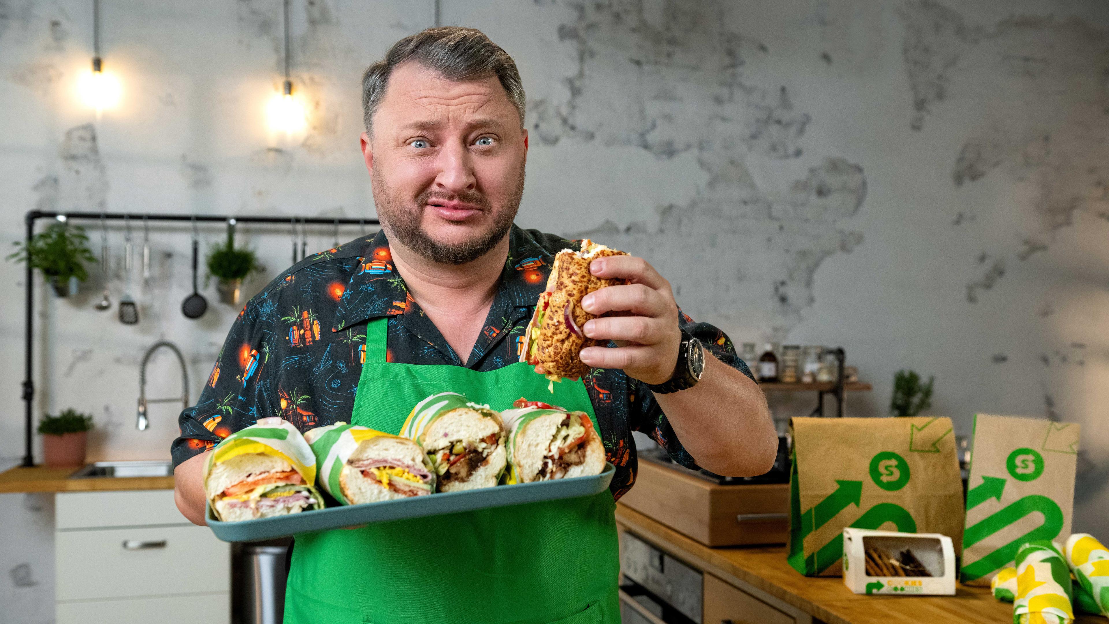 Sebastian Lege steht an einer Arbeitsfläche und hält in der einen Hand ein Tablett mit Sandwiches von Subway. In der anderen Hand hält er ein angebissenes Sandwich. Dabei verzieht er sein Gesicht.