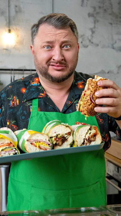 Sebastian Lege steht an einer Arbeitsfläche und hält in der einen Hand ein Tablett mit Sandwiches von Subway. In der anderen Hand hält er ein angebissenes Sandwich. Dabei verzieht er sein Gesicht.