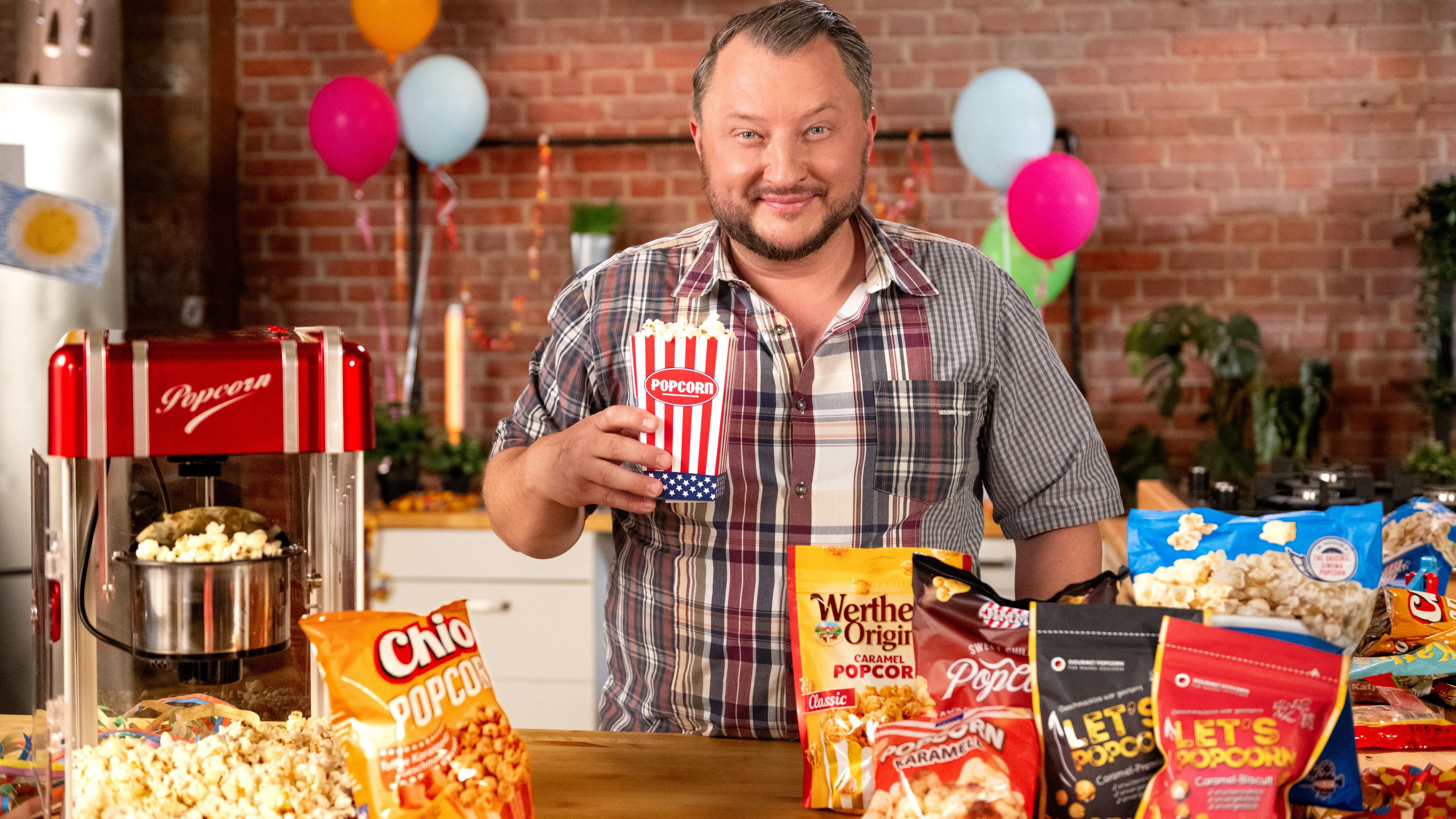 Sebastian Lege hält eine Popcorn-Tüte in der Hand, vor ihm liegen verschiedene Popcorn-Packungen auf einem Tisch.