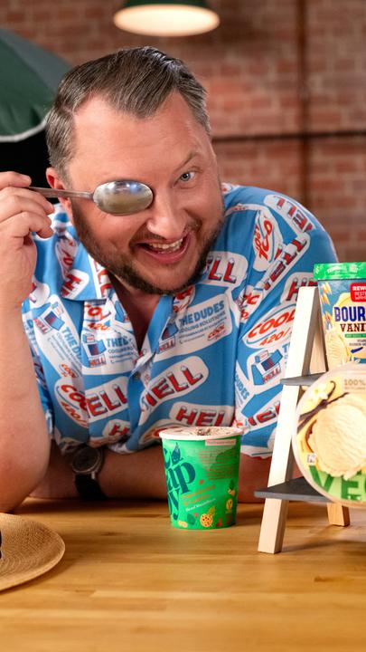 Sebastian Lege hält sich einen Löffel vor das rechte Auge, neben ihm stehen verschiedene vegane Packungen mit Eis.