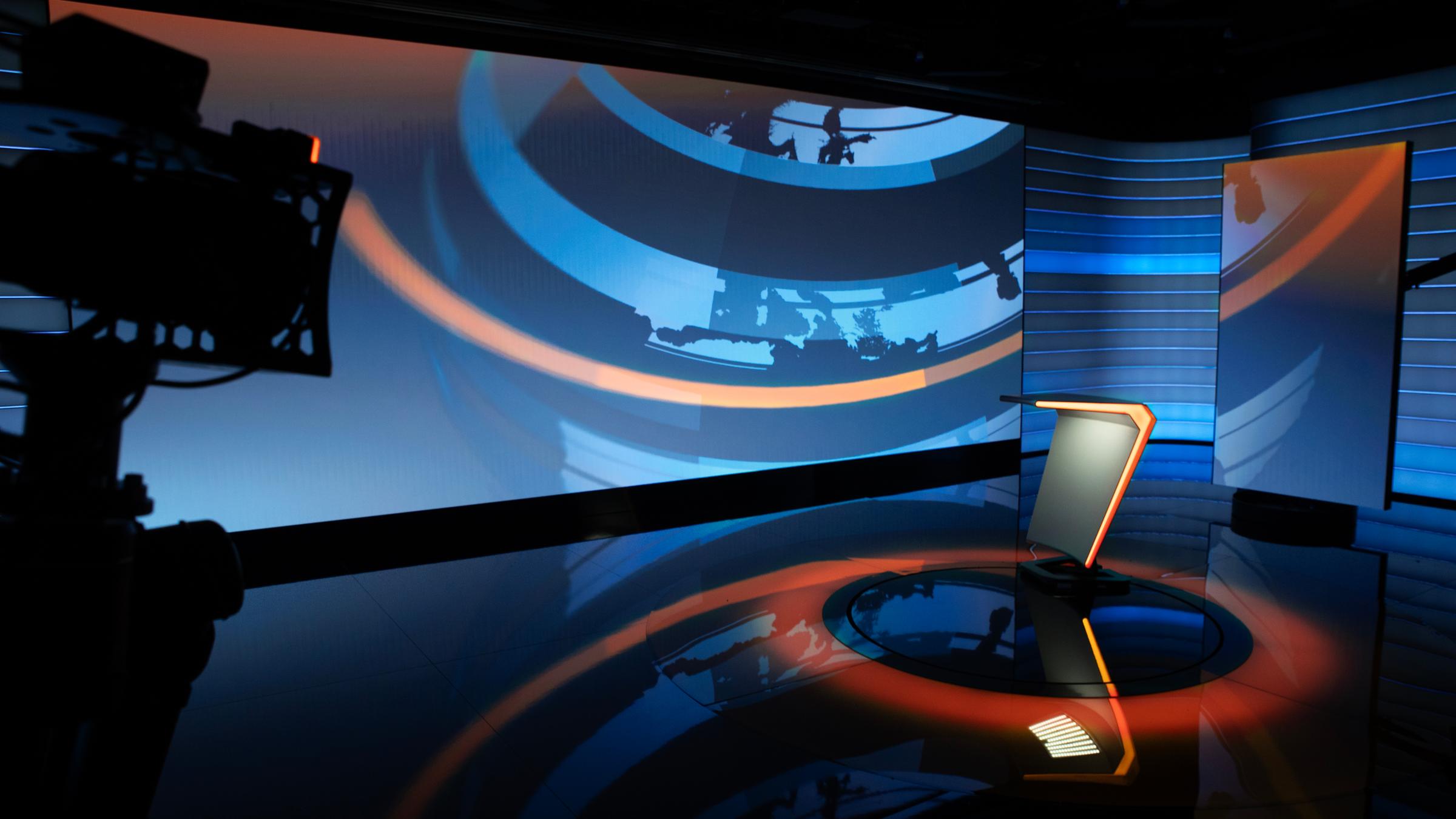 Im neuen Online-Nachrichtenstudio steht ein Moderationstisch mit orangener Kante und einem grafischen Hintergrund in blauen Schattierungen und orange. 