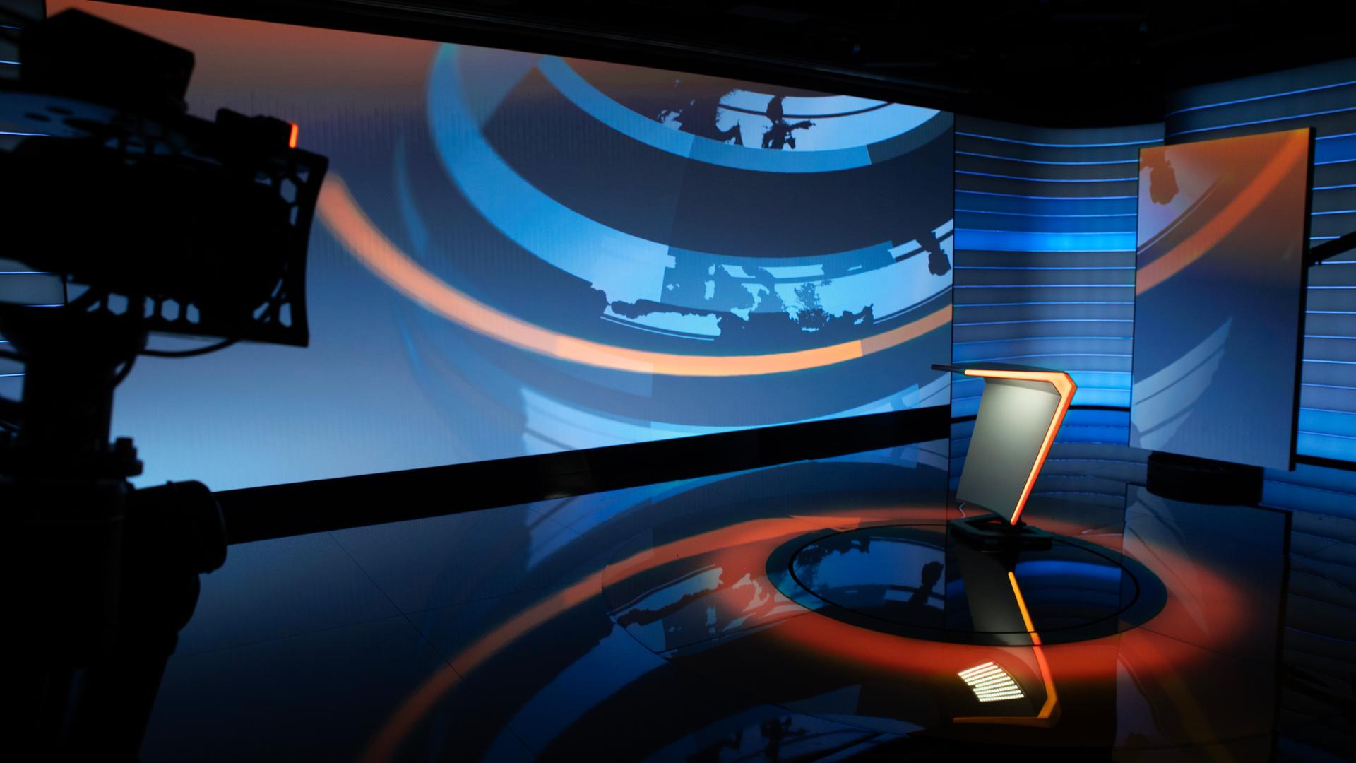 Im neuen Online-Nachrichtenstudio steht ein Moderationstisch mit orangener Kante und einem grafischen Hintergrund in blauen Schattierungen und orange. 