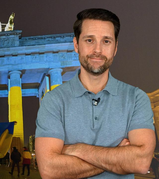 Im Hintergrund zu erkennen ist das Brandenburger Tor in Berlin, davor Menschen mit Ukraine-Flaggen. Rechts im Bild zu sehen ist der Host Mirko Drotschmann.