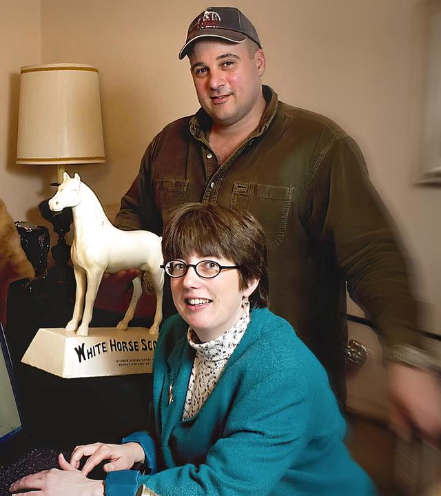 Ina und David Steiner befinden sich in einem Raum, Ina sitzt vor einem Laptop, David steht hinter ihr. Auf dem Tisch steht eine weiße Pferdefigur, an der Seite auf einer Schneiderpuppe hängt eine braune Pelzjacke.