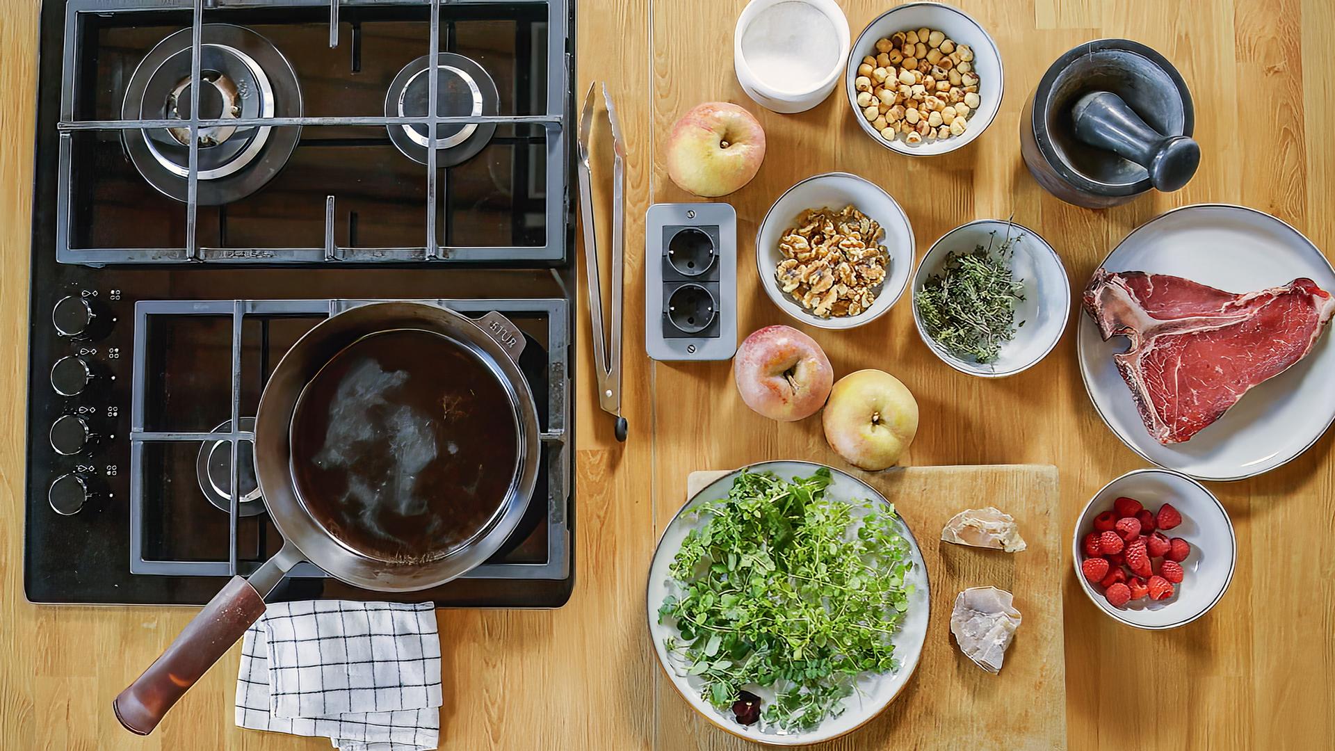 Auf einer Holzplatte sind zwei Kochfelder, auf einem steht eine Pfanne. Daneben sind die Zutaten zu einem Gericht auf Tellern und in Schüsseln bereitgestellt.