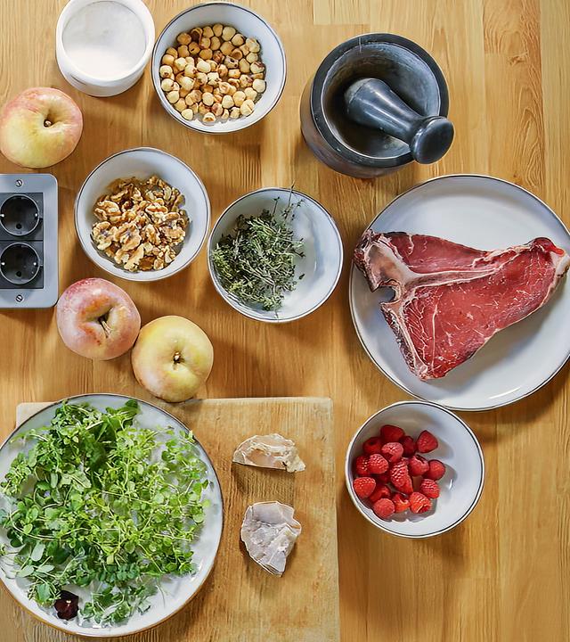 Auf einer Holzplatte sind zwei Kochfelder, auf einem steht eine Pfanne. Daneben sind die Zutaten zu einem Gericht auf Tellern und in Schüsseln bereitgestellt.