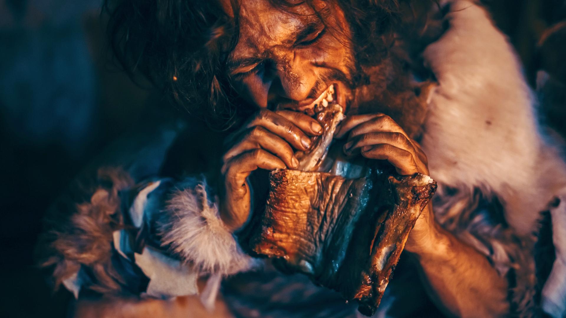 Nahaufnahme, ein Höhlenmensch beißt von einem großen Stück Fleisch ab.