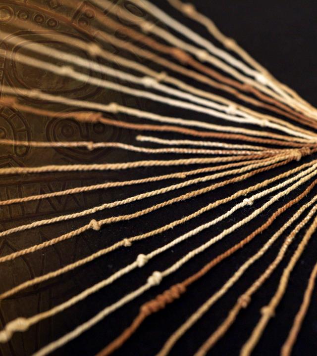 Die Nahaufnahme eines Knotenbändchens der Inka liegt vor schwarzem Hintergrund. An der Seite überlagert das Muster einer Sonnenscheibe der Inka das Foto.