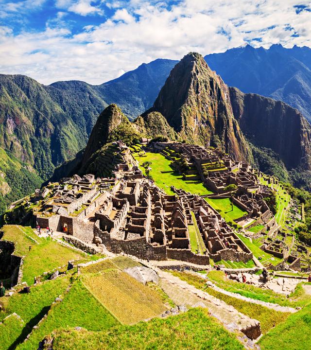 Eine Luftaufnahme des Machu Picchu mit Inkaruinen liegt im Sonnenschein unter blauem Himmel. An der Seite überlagert das Muster einer Sonnenscheibe der Inka das Landschaftspanorama.