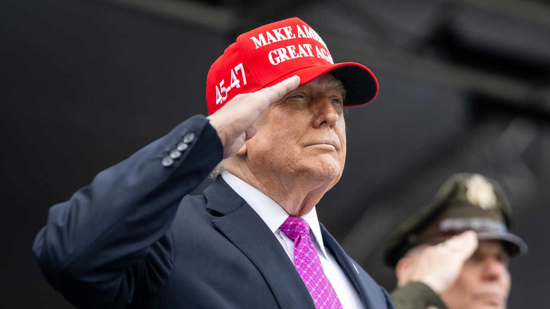 Donald Trump trägt eine rote "Make America Great Again"-Kappe und salutiert. Im Hintergrund steht ein weiterer salutierender Mann, der eine US-amerikanische Militäruniform trägt.