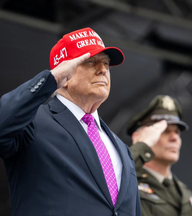 Donald Trump trägt eine rote "Make America Great Again"-Kappe und salutiert. Im Hintergrund steht ein weiterer salutierender Mann, der eine US-amerikanische Militäruniform trägt.
