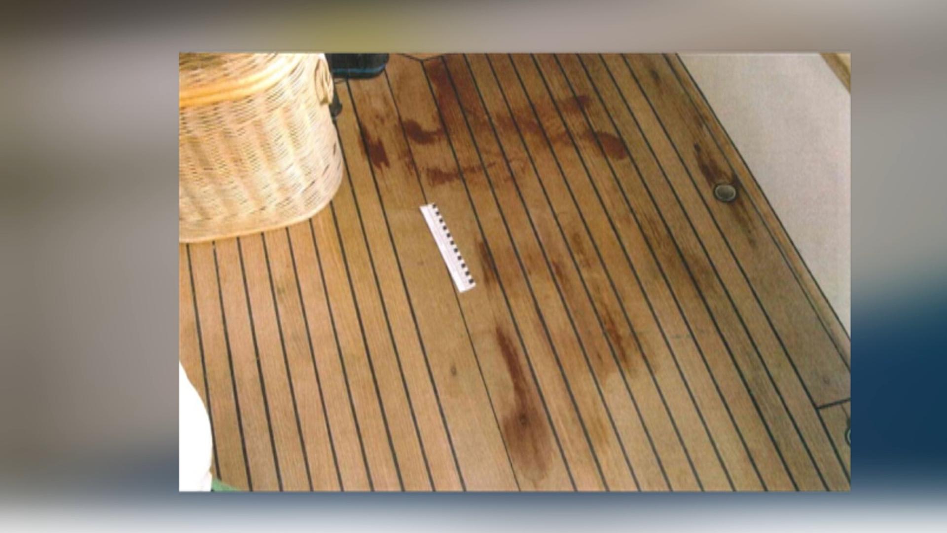 Auf einem braunen Holzboden sind rote Flecken sichtbar. Neben einem beigen Korb liegt ein weiß-schwarzes Lineal, das die Größe der Flecken misst.