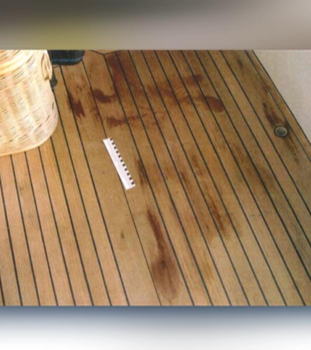 Auf einem braunen Holzboden sind rote Flecken sichtbar. Neben einem beigen Korb liegt ein weiß-schwarzes Lineal, das die Größe der Flecken misst.